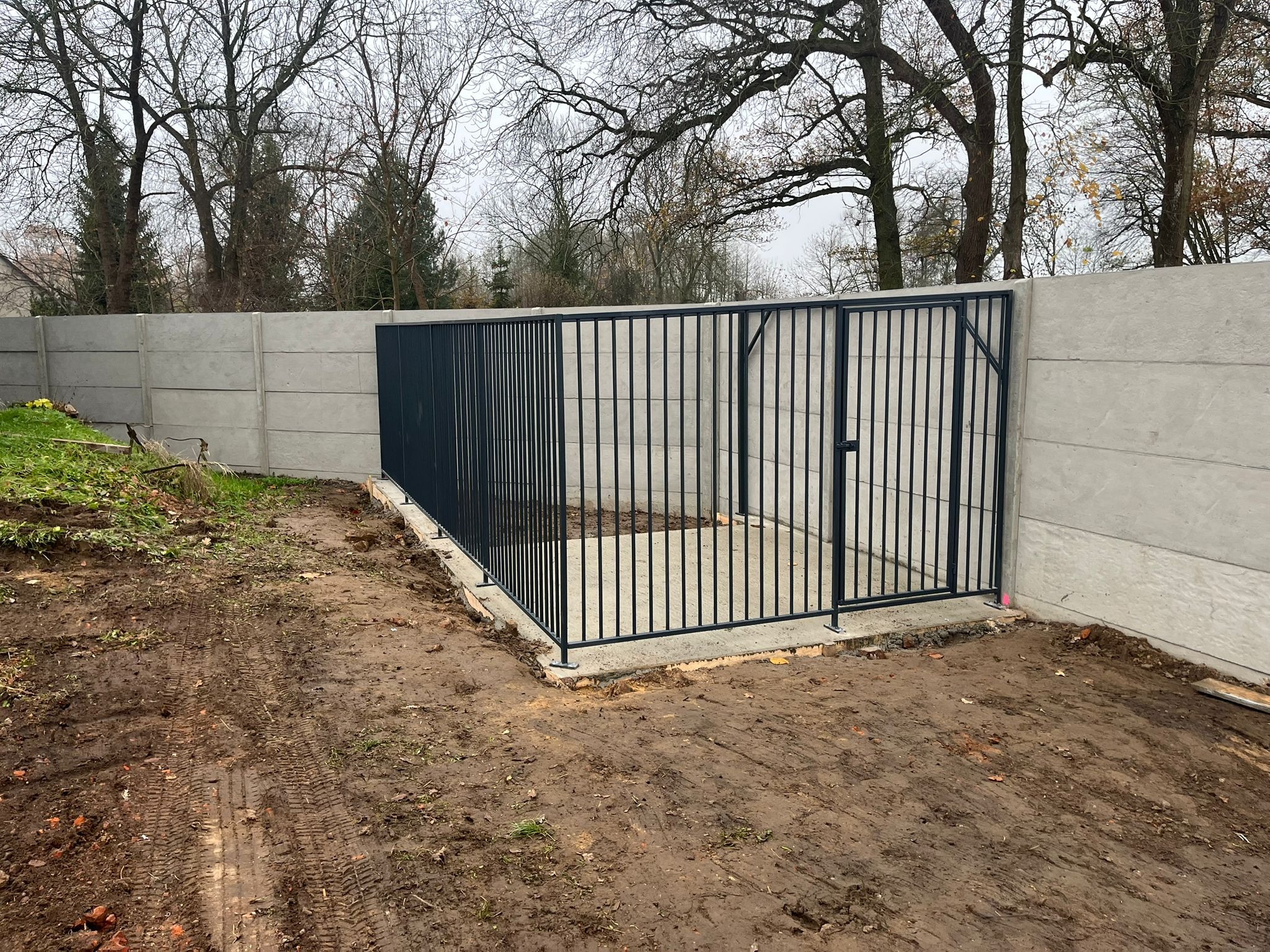 Nowoczesna, metalowa brama wjazdowa z pionowymi prętami, osadzona w betonowym fundamencie, na tle betonowego ogrodzenia panelowego. Surowy krajobraz, pochmurne niebo.