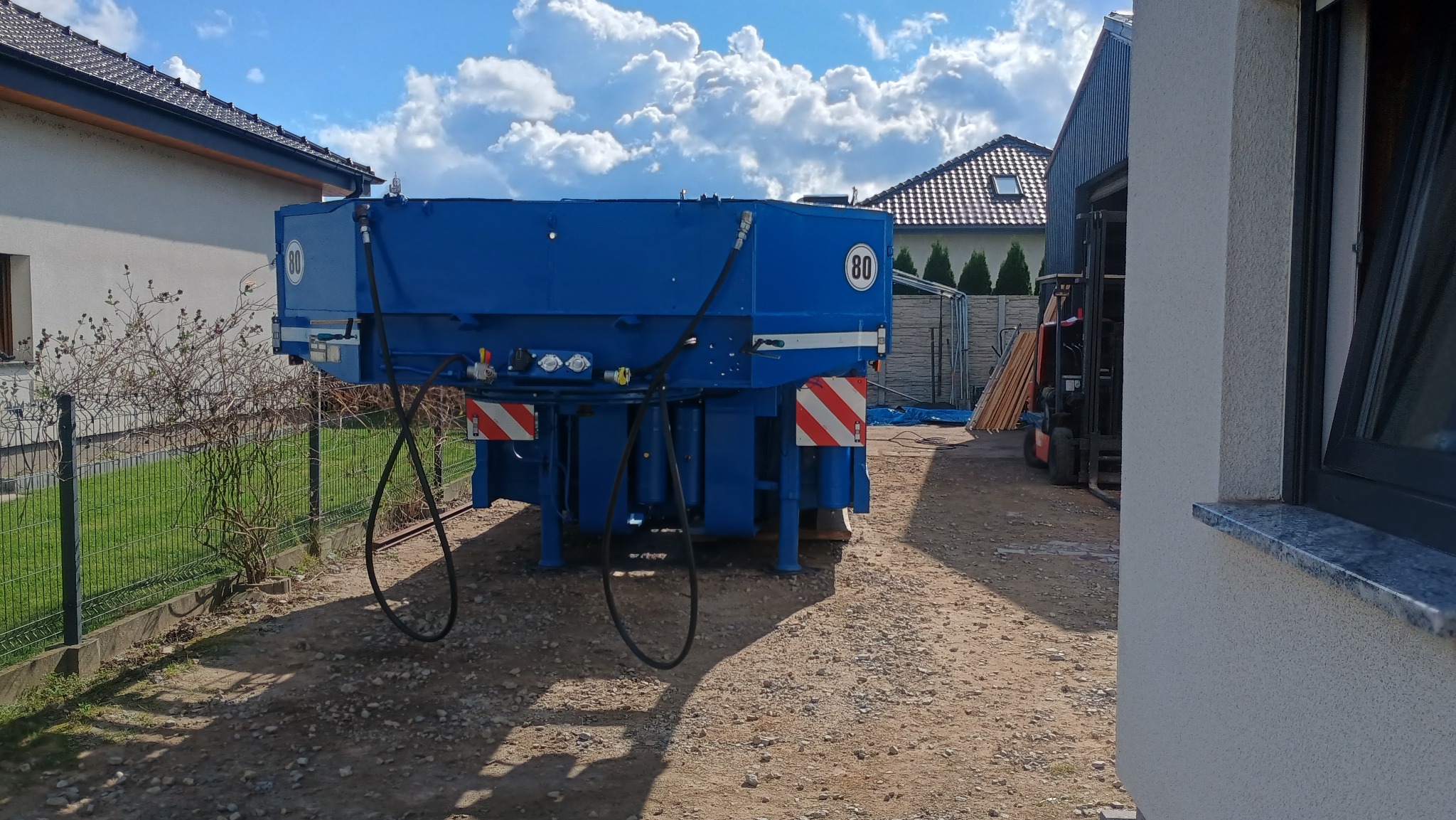 Niebieska, metalowa konstrukcja z oznaczeniem prędkości 80 km/h, stojąca na żwirowym podłożu, w tle budynki mieszkalne i wózek widłowy.