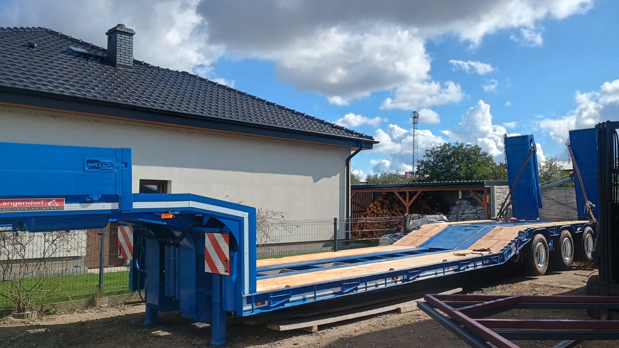 Niebieska, niskopodwoziowa przyczepa Langendorf w Rychtalu, z drewnianą podłogą, na tle domu i błękitnego nieba z chmurami. Widoczne elementy konstrukcyjne.