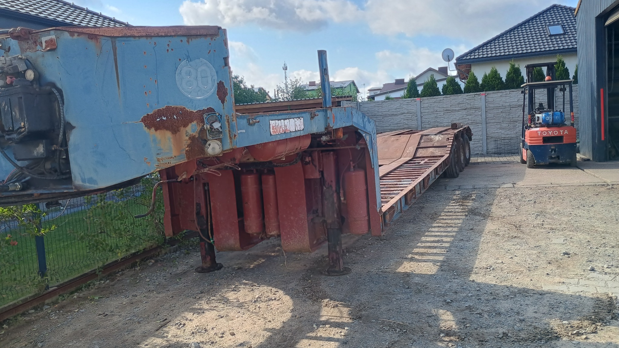 Niskozawieszona, skorodowana przyczepa z widocznym podwoziem i elementami hydraulicznymi, obok wózek widłowy Toyota, na tle zabudowań.