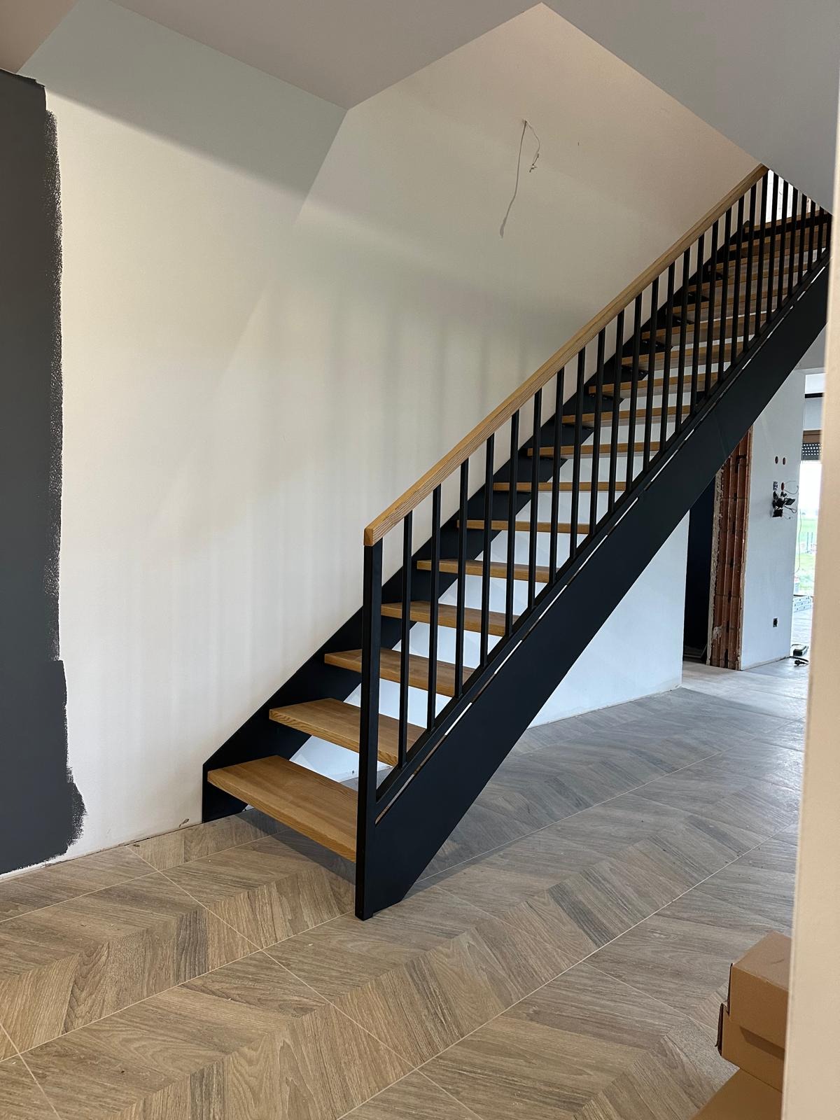 Nowoczesne schody z drewnianymi stopniami i czarną metalową konstrukcją, widoczne na tle jasnego wnętrza z częściowo pomalowaną ścianą na szaro.