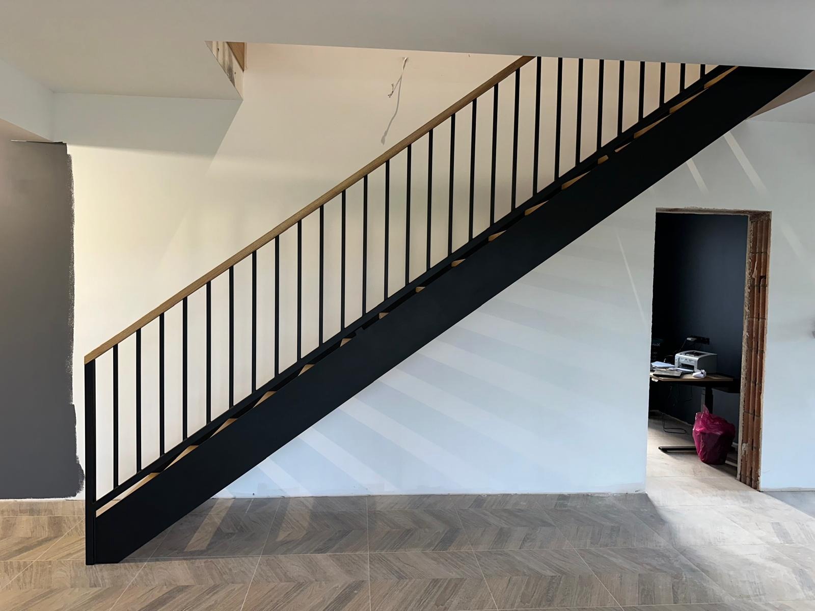 Nowoczesne schody metalowe z drewnianą poręczą, czarne barierki, widok z dołu na tle białej ściany z szarym fragmentem i wzorzystej podłogi. Minimalistyczny design.