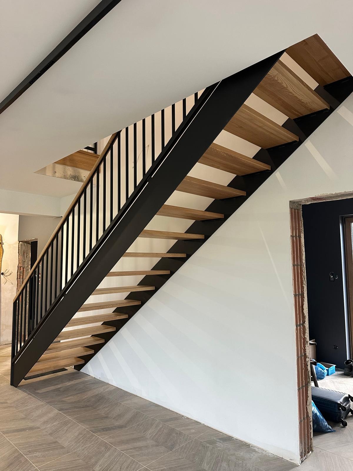 Nowoczesne schody z drewnianymi stopniami i czarną, metalową konstrukcją, widoczne z dołu, z fragmentem podłogi w jodełkę i surowym otworem drzwiowym w tle.