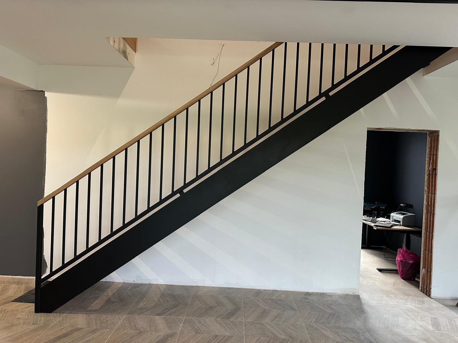 Nowoczesne schody z czarną metalową konstrukcją i drewnianą poręczą, widoczne na tle białej ściany i podłogi z płytek w jodełkę. Minimalistyczny design.