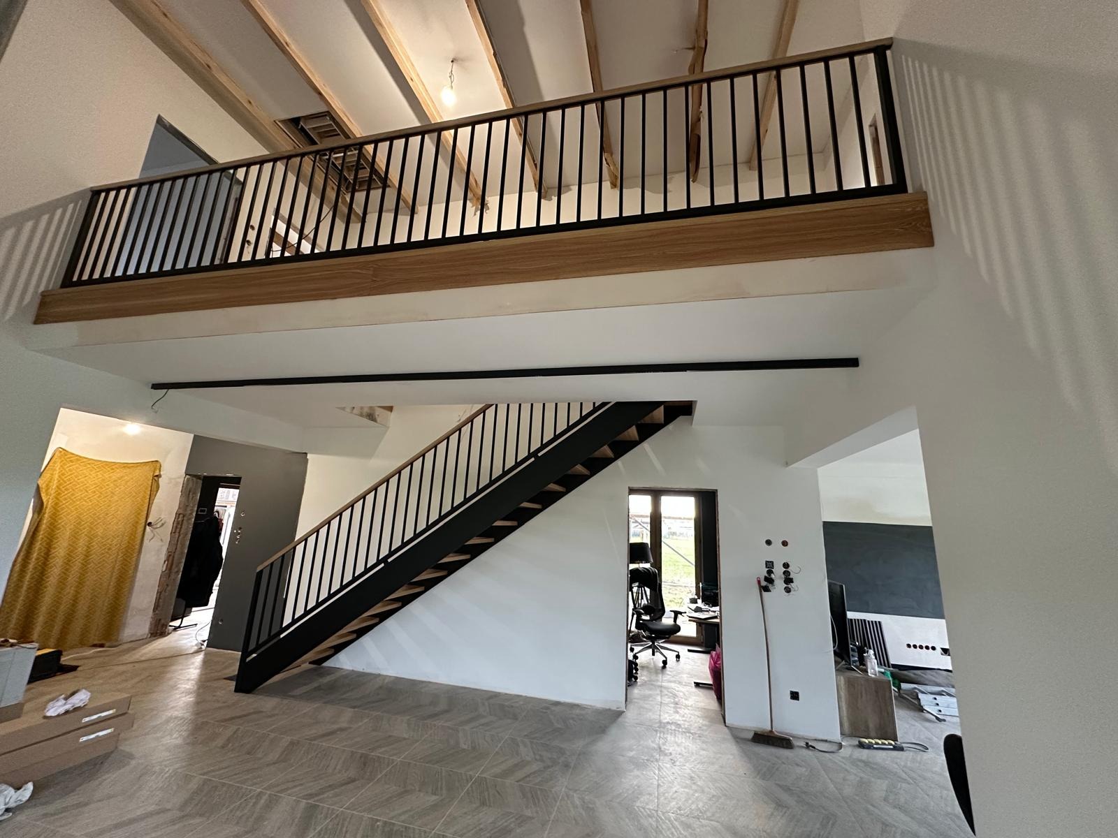 Nowoczesne schody metalowe z drewnianymi stopniami i czarną balustradą, prowadzące na antresolę z drewniano-metalową balustradą w jasnym, przestronnym wnętrzu.