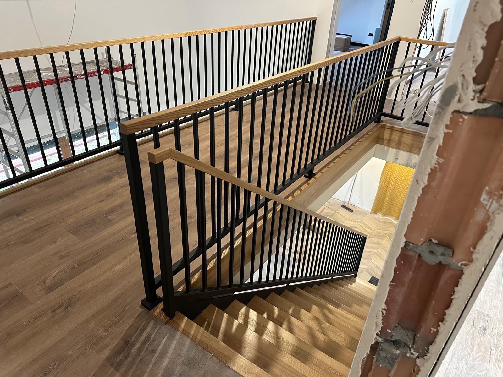 Nowoczesne schody z drewnianymi stopniami i czarną, metalową balustradą z drewnianym poręczą. Widok z góry na dół, z elementami wykończeniowymi w tle.