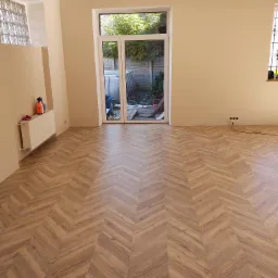 Nowo ułożona podłoga z paneli w jodełkę w jasnym, odświeżonym pokoju, z widocznymi gniazdkami elektrycznymi bez osprzętu i grzejnikiem z akcesoriami malarskimi.