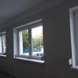 Wnętrze po malowaniu na biało, widoczne trzy okna z białymi ramami i parapetami, surowa podłoga, brak mebli, ściany świeżo pomalowane.