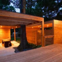 Nowoczesna drewniana pergola z wbudowanym drzewem, oświetlona punktowymi lampami, połączona z drewnianym tarasem i budynkiem z drewnianą elewacją