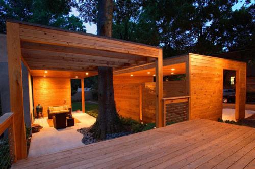 Nowoczesna drewniana pergola z wbudowanym drzewem, oświetlona punktowymi lampami, połączona z drewnianym tarasem i budynkiem z drewnianą elewacją