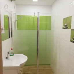 Nowoczesna łazienka z zielonymi akcentami: kabina prysznicowa z matową szybą, biała umywalka z szafką, drewnopodobne płytki na podłodze, oraz dekoracyjne płytki z motywem kamieni zen na ścianie.