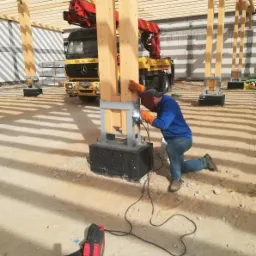 Spawanie stalowej podstawy drewnianego słupa hali, z dźwigiem samochodowym w tle, widoczny spawacz w niebieskiej bluzie i masce ochronnej.