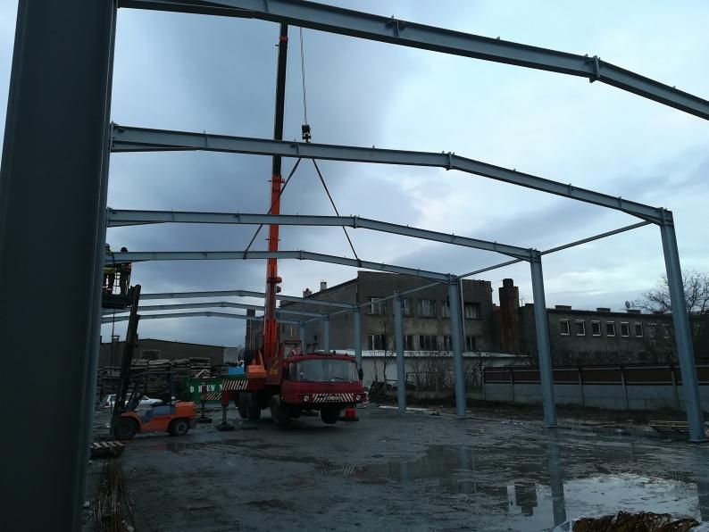 Montaż szkieletu stalowej hali z użyciem dźwigu, widoczny wózek widłowy i pracownicy na podnośniku, tło stanowią budynki przemysłowe pod pochmurnym niebem.