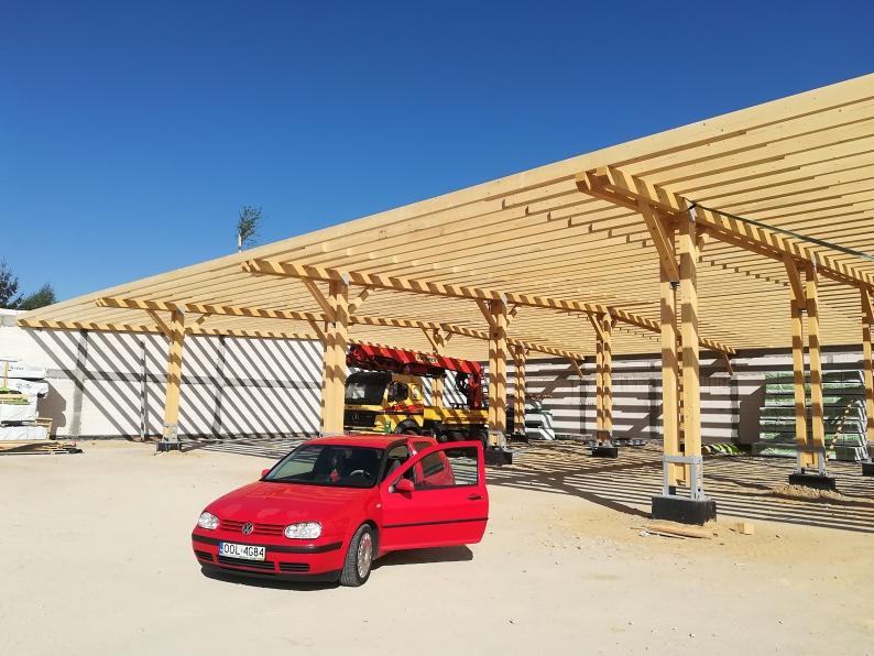 Drewniana konstrukcja zadaszenia w trakcie budowy, widoczny czerwony samochód osobowy i dźwig w tle.