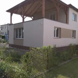 Dwukondygnacyjny budynek po remoncie elewacji w odcieniach beżu i brązu, z drewnianą konstrukcją zadaszenia nad balkonem na pierwszym piętrze, widoczny z poziomu gruntu przez zarośla i siatkę...