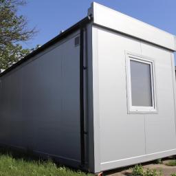 KAMP KONTENER S.C. PRODUCENT KONTENERÓW - Szary, modułowy pawilon z jednym oknem, widoczny fragment rynny, trawa i betonowe podłoże.