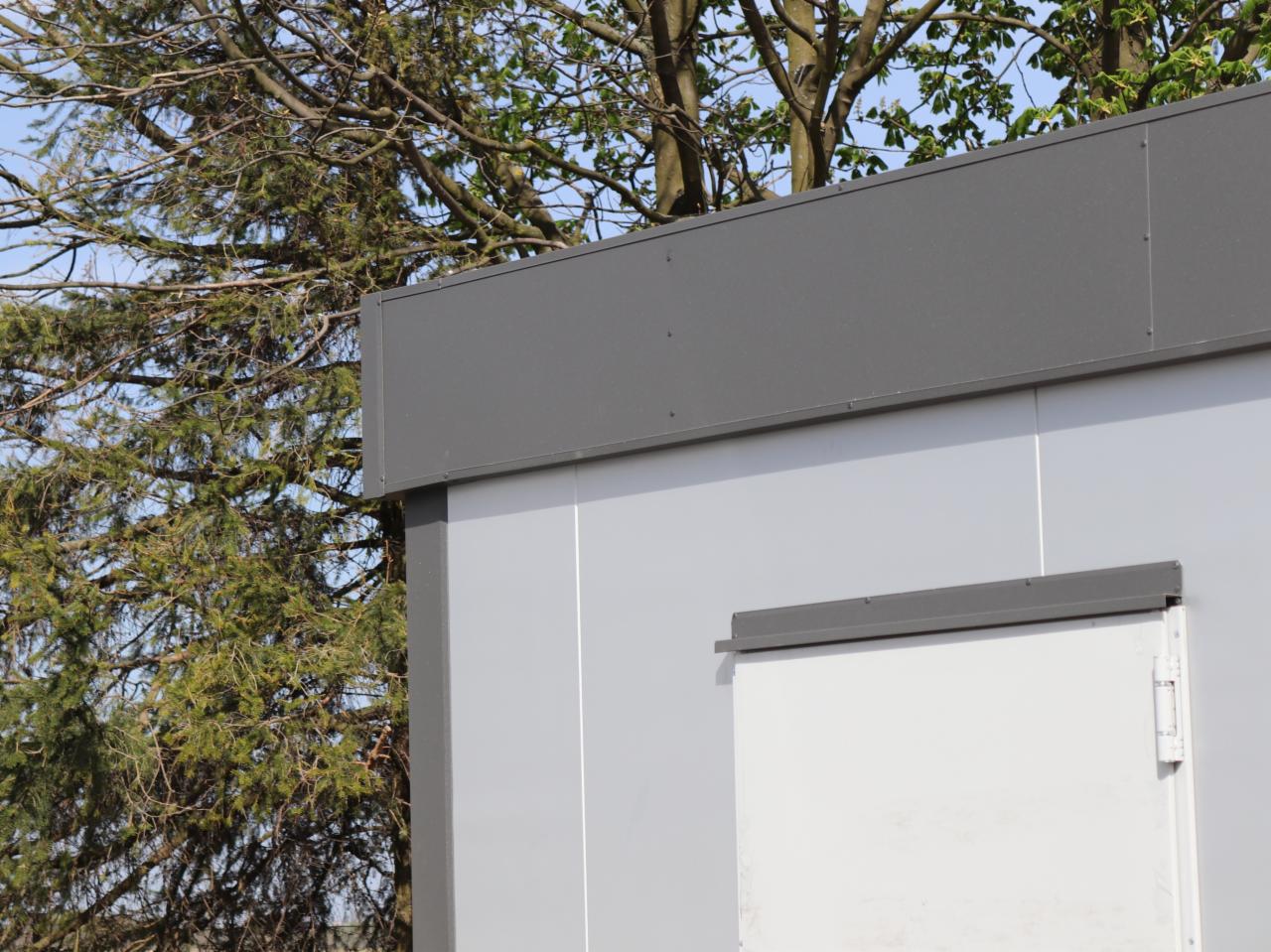 Szary, modułowy budynek z białą, zamkniętą furtką, widoczny na tle drzew