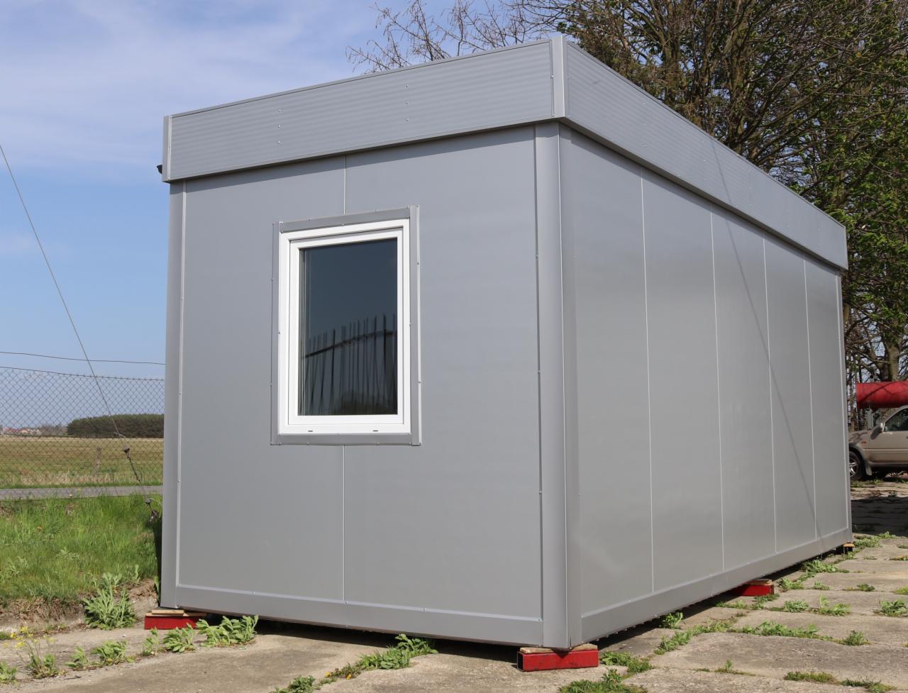 Szary kontener biurowy z białym oknem, ustawiony na czerwonych podkładkach, widok z zewnątrz na tle trawy i drzew.