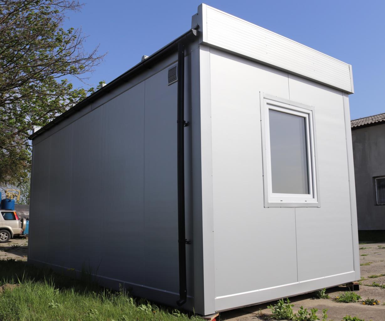 Szary, modułowy pawilon z jednym oknem, widoczny fragment rynny, trawa i betonowe podłoże.