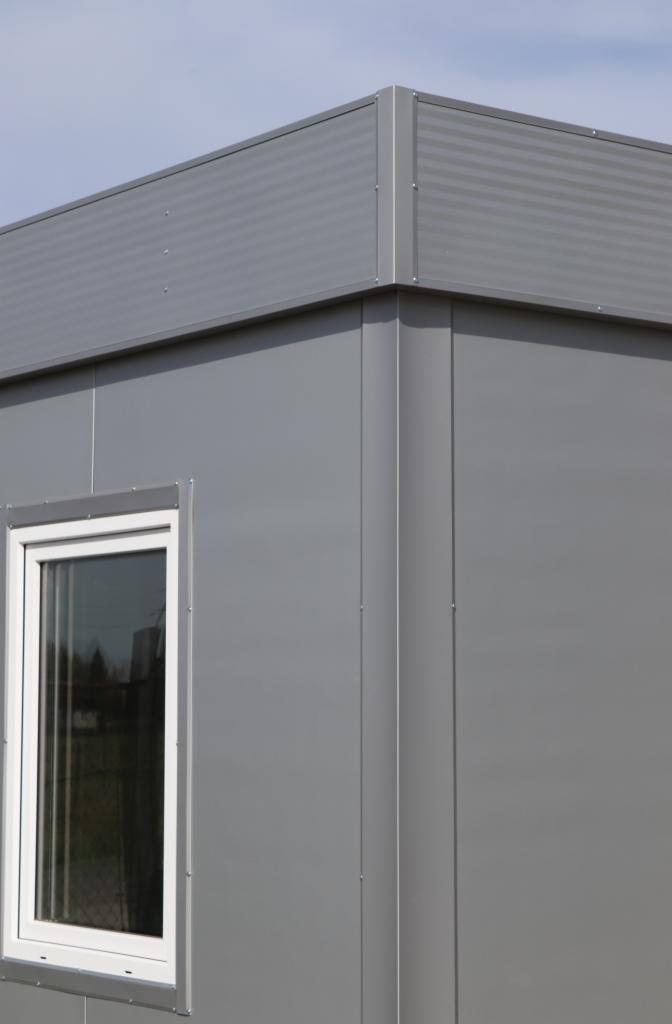 Szary, modułowy budynek z białym oknem, widok z zewnątrz na narożnik i fragment dachu.