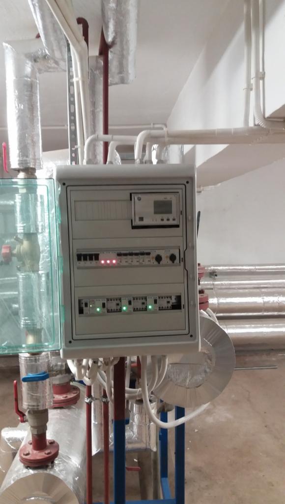 Szara szafa sterownicza z widocznymi przewodami, zabezpieczeniami i wskaźnikami LED, umieszczona w pomieszczeniu technicznym z rurami w izolacji termicznej.