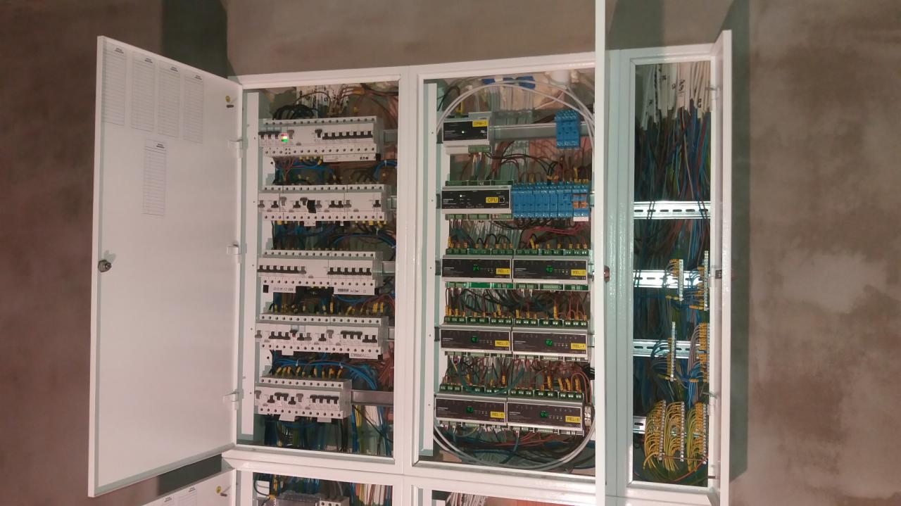 Rozdzielnica elektryczna z otwartymi drzwiami, widoczne liczne przewody, bezpieczniki i inne komponenty, zamontowana na surowej ścianie w trakcie instalacji.