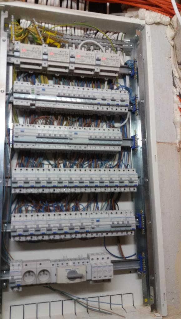 Otwarta skrzynka elektryczna z licznymi bezpiecznikami, przewodami i gniazdkami, zamontowana na ścianie z widoczną pianką montażową.