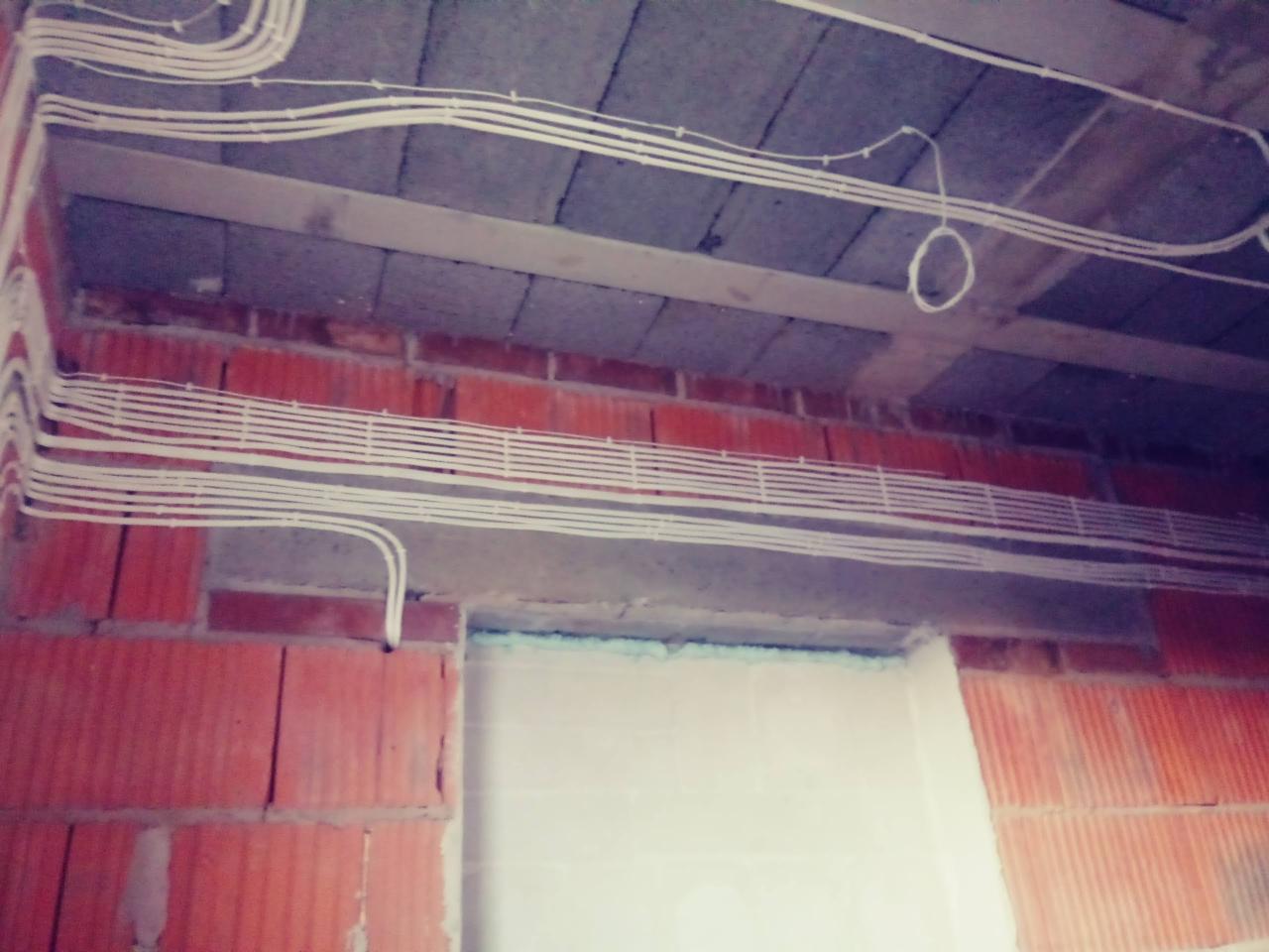 Surowe wnętrze w trakcie budowy, widoczne liczne białe przewody elektryczne ułożone równolegle pod sufitem z betonowych płyt, ściany z czerwonej cegły.