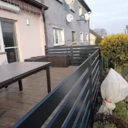 Nowoczesna, pozioma, grafitowa balustrada na tarasie z drewnopodobną podłogą, widok na dom z antenami satelitarnymi i roślinność ogrodową, w tym zabezpieczony krzew.