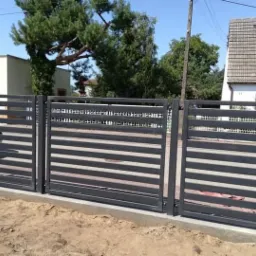 Nowoczesna, przesuwna brama posesyjna z poziomymi profilami w kolorze grafitowym, świeżo zamontowana na betonowej podmurówce z widocznymi śladami prac ziemnych.