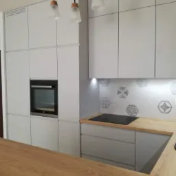 Nowoczesna, jasnoszara kuchnia z drewnianym blatem, heksagonalnymi płytkami dekoracyjnymi nad płytą indukcyjną oraz trzema lampami wiszącymi z drewnianymi akcentami.