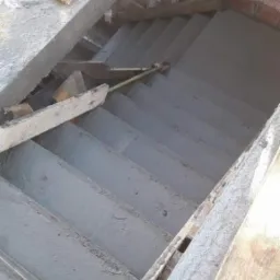 Surowe, betonowe schody w trakcie budowy wewnątrz murowanego budynku, widoczne drewniane elementy szalunku i ceglana ściana w tle.