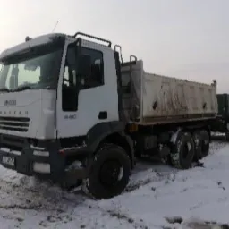 Biała ciężarówka Iveco Tracker 440 z przyczepą wywrotką, częściowo pokryta śniegiem, stoi na zasypanym śniegiem terenie.