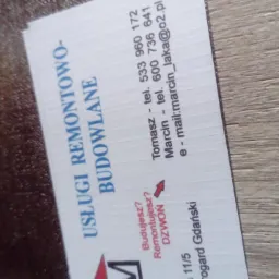 Wizytówka firmy remontowo-budowlanej leżąca na drewnopodobnej powierzchni, z widocznymi danymi kontaktowymi i hasłem 'Budujesz? Remontujesz? DZWOŃ'.