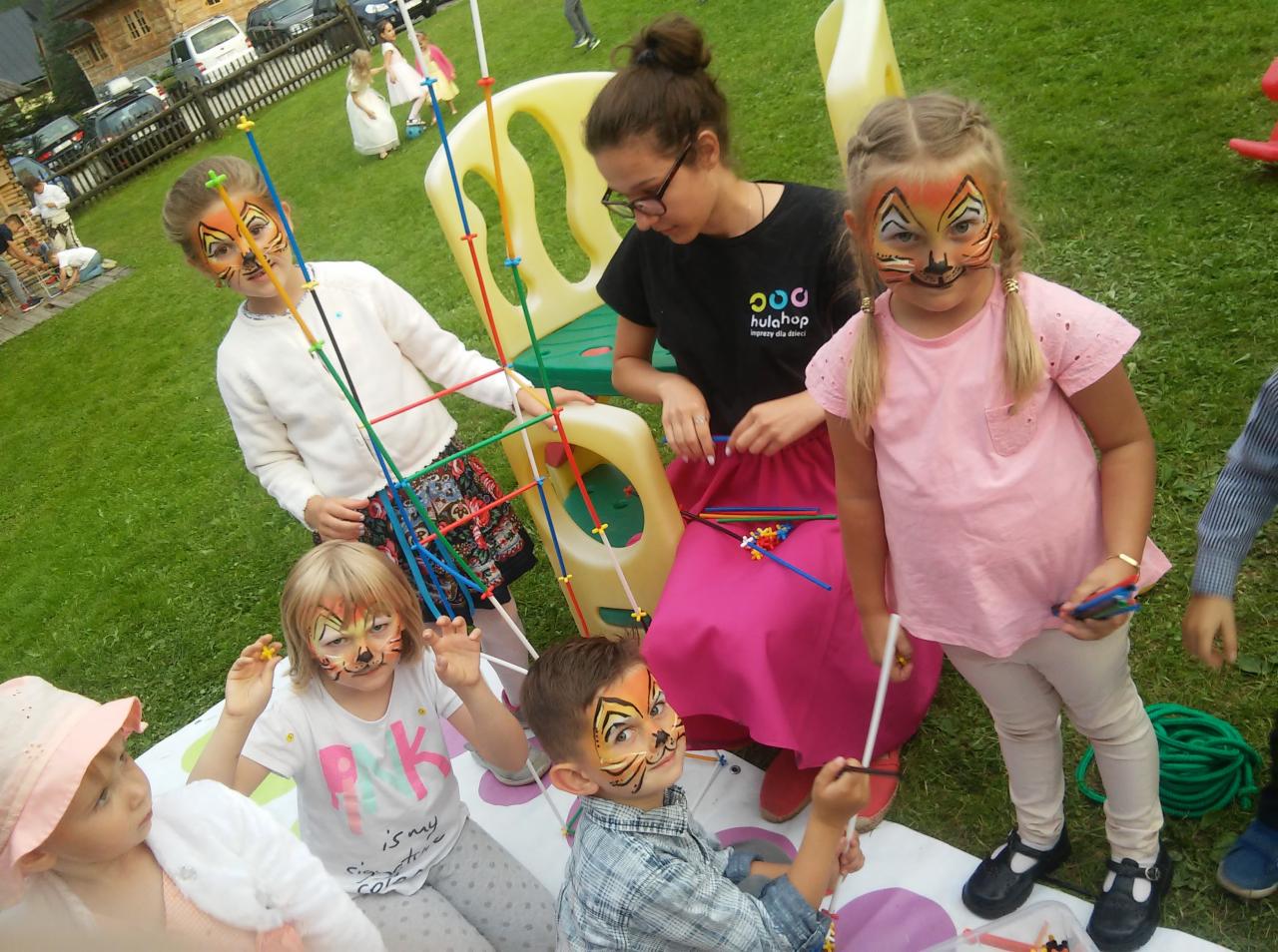 Grupa dzieci z pomalowanymi twarzami w motywy zwierzęce, bawiących się kolorowymi patyczkami i elementami konstrukcyjnymi pod okiem kobiety w czarnej koszulce z logo firmy eventowej, na trawniku...