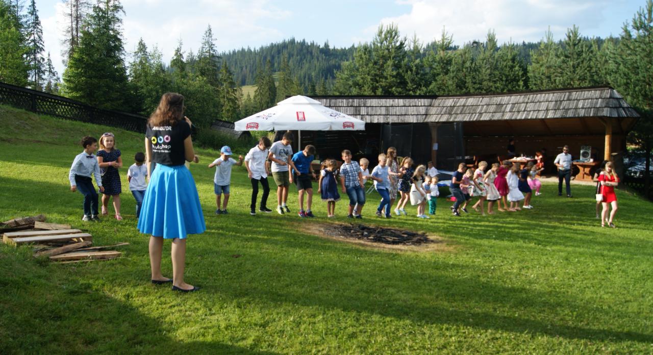 Grupa dzieci ustawiona w rzędzie na zielonej trawie, prowadzona przez osobę w niebieskiej spódnicy i czarnej koszulce z napisem 'Animator', w tle zadaszona konstrukcja, parasol i drzewa.