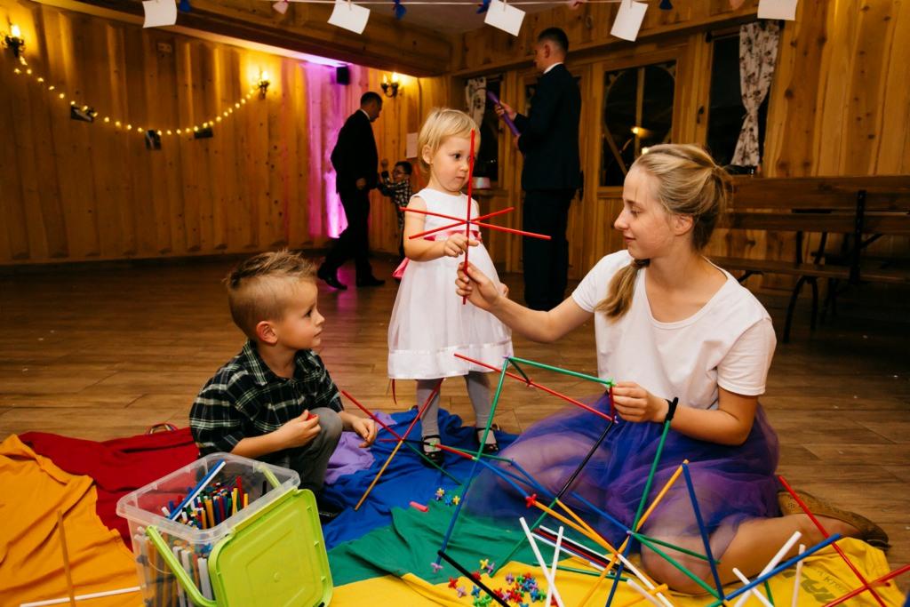 Animator pomaga dzieciom w budowaniu konstrukcji z kolorowych patyczków w sali z drewnianymi ścianami i dekoracjami świetlnymi.