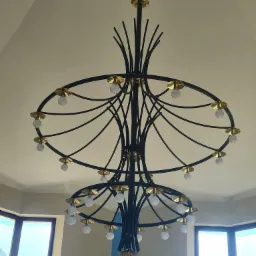 Dwupoziomowy żyrandol z czarnymi ramionami i złotymi detalami, widok z dołu na tle kremowego sufitu i okien.