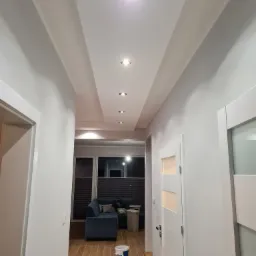 Białe ściany i sufit w przedpokoju z wbudowanymi lampami punktowymi, widoczny fragment salonu z niebieską kanapą oraz akcesoria malarskie przy drzwiach.