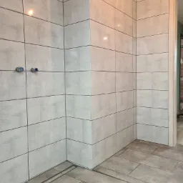 Wykończona łazienka z szarymi, kwadratowymi płytkami na ścianach i podłodze, widoczny odpływ liniowy, instalacja wodna przygotowana do podłączenia armatury.