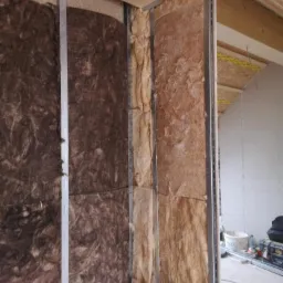 Montaż izolacji z wełny mineralnej między metalowymi profilami konstrukcyjnymi ściany działowej, widoczne fragmenty płyty OSB i narzędzia w tle.