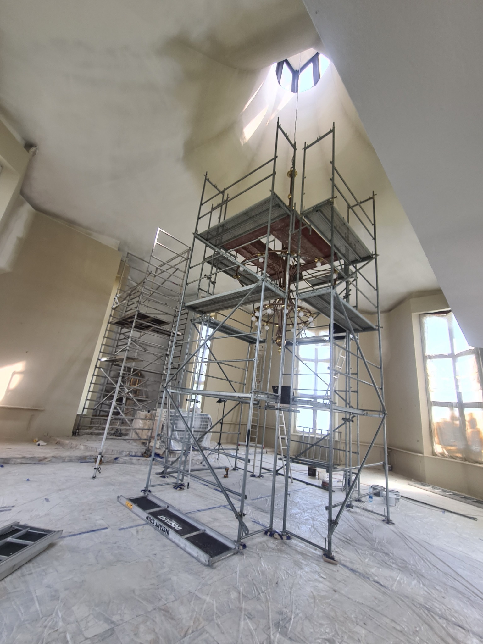 Wysokie rusztowanie w przestronnym wnętrzu z oknami i świetlikiem, podłoże zabezpieczone folią malarską, widoczne ślady prac na ścianach.