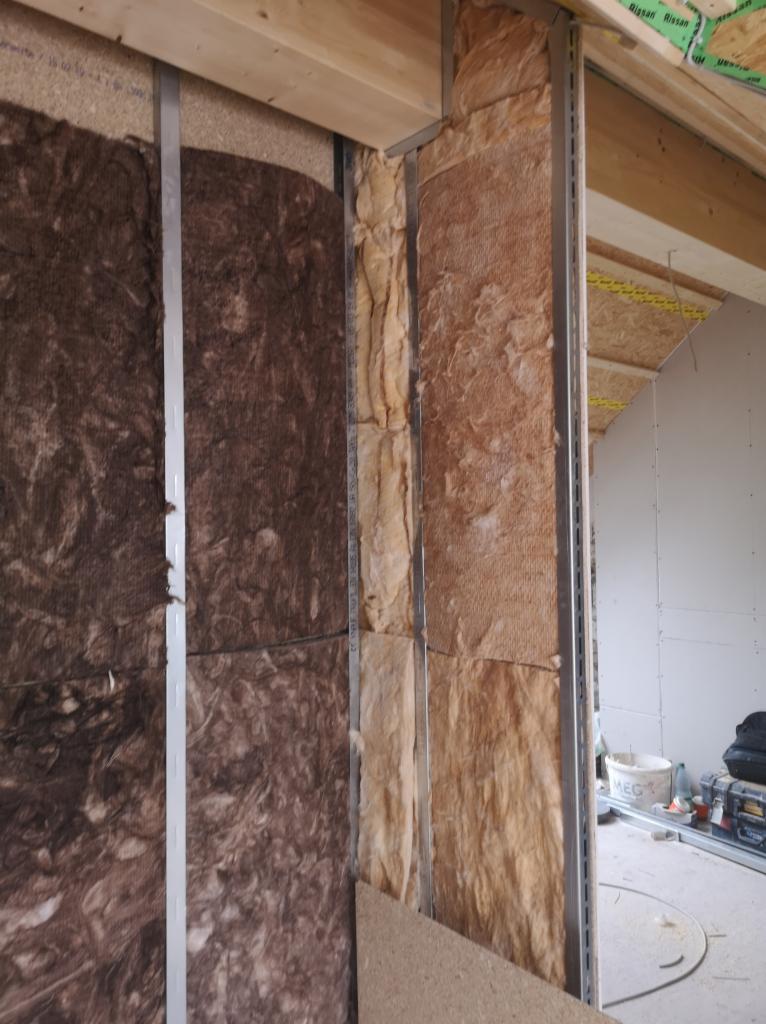 Montaż izolacji z wełny mineralnej między metalowymi profilami konstrukcyjnymi ściany działowej, widoczne fragmenty płyty OSB i narzędzia w tle.