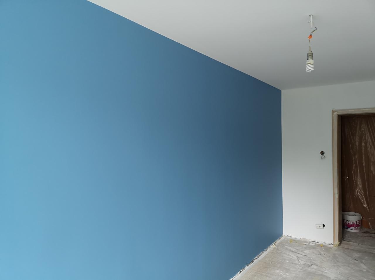 Świeżo pomalowana na niebiesko ściana w pokoju, widoczny pojemnik z farbą RAPID przy drzwiach, sufit biały, niedokończone wykończenie podłogi.