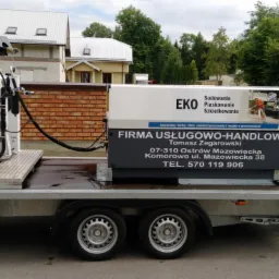 Mobilny zestaw do obróbki strumieniowo-ściernej na przyczepie, z widocznym zbiornikiem, przewodami i tablicą informacyjną firmy z Ostrowi Mazowieckiej, na tle zabudowań mieszkalnych i zaparkowanego...