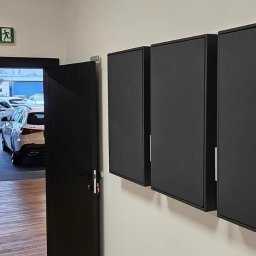 Safety House - Trzy czarne szafki wiszące na białej ścianie, widoczne przy wejściu do salonu samochodowego z zaparkowanymi autami.