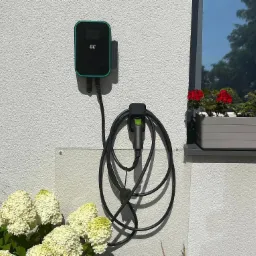 Na elewacji domu zamontowana stacja ładowania pojazdów elektrycznych marki GC z podpiętym kablem, obok kwitnące hortensje i skrzynka z czerwonymi pelargoniami w oknie.