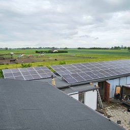 Safety House - Panele fotowoltaiczne na dachu budynku gospodarczego na tle łąki z pasącymi się zwierzętami, widoczny samochód dostawczy i drabina.