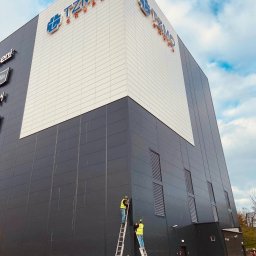 Safety House - Dwóch pracowników na drabinach montuje lub naprawia elementy elewacji budynku z logo TZMO Grupa, na tle błękitnego nieba z chmurami.