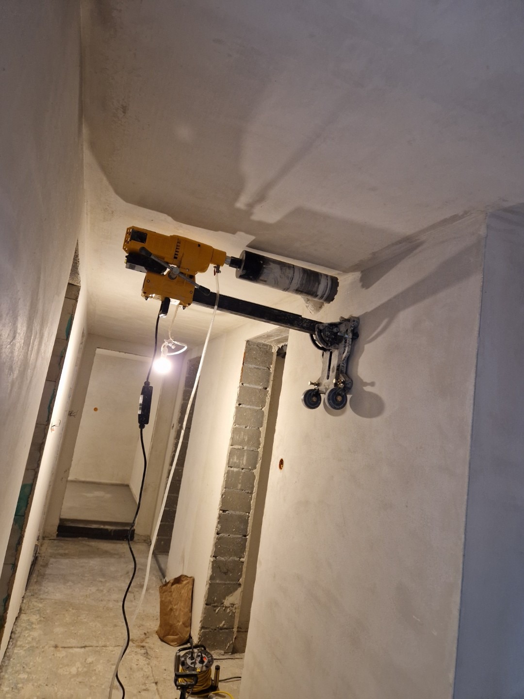Żółta wiertnica diamentowa zamocowana do ściany i sufitu w surowym wnętrzu, wiercąca otwór w suficie. Widoczne przewody, lampa robocza i częściowo wykończone ściany.